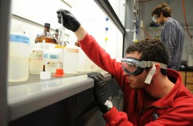 Student in a red jacket and safety gear kneeling on the ground and pipetting into a graduated cylinder